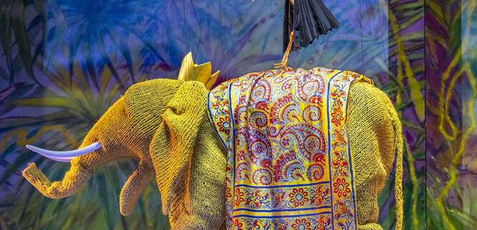 A colorful elephant with a magnificent saddle pattern stands in the foreground. A stork balances on its back. | © Bernhard Müller