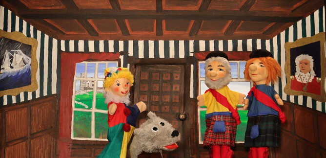 Three hand puppets are standing in a stylish room with wood paneling. A fur dog lies in front of them, while paintings hang on the walls. | © Friedburger Puppenbühne
