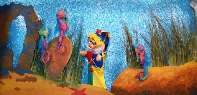 A colorful underwater world with a puppeteer and colorful seahorses. In the background, seaweed and sandy underwater landscapes can be seen. | © Friedburger Puppenbühne