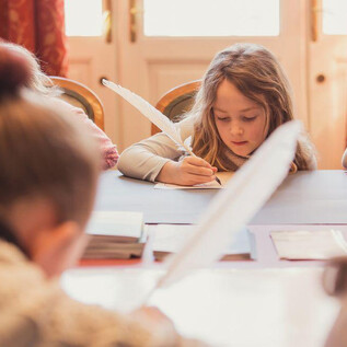 A girl is writing at a table, surrounded by other children. On the table are a pen, paper, and school supplies. | © Eva trifft