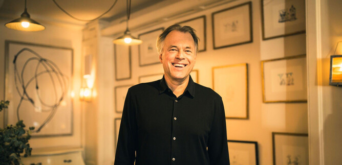 A smiling man stands in a stylish room with illuminated walls. Framed pictures hang in the background, creating a creative atmosphere. | © Mina Esfandiari