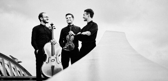 Drei Musiker stehen auf einer Treppe und lächeln. Einer hält ein Cello, während die anderen eine Violine und ein weiteres Instrument halten. | © Kaupo Kikkas