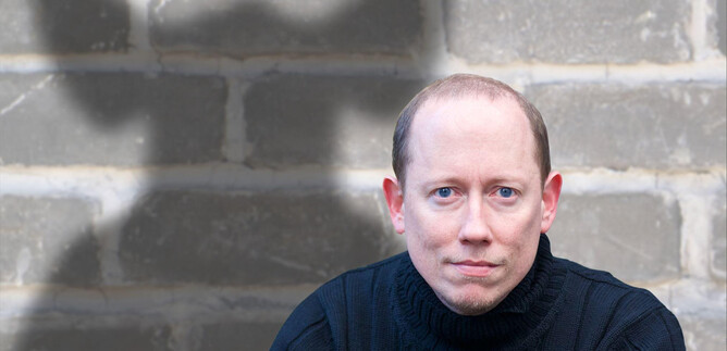 A man is sitting in front of a gray wall with a serious expression. In the background, a shadow is visible that has an interesting shape. | © ARGEkultur
