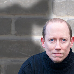 A man is sitting in front of a gray wall with a serious expression. In the background, a shadow is visible that has an interesting shape. | © ARGEkultur