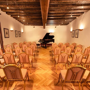 An elegant room with wooden floors and ceiling beams. In the center stands a piano, surrounded by many chairs for the audience. | © Agentur Orpheus