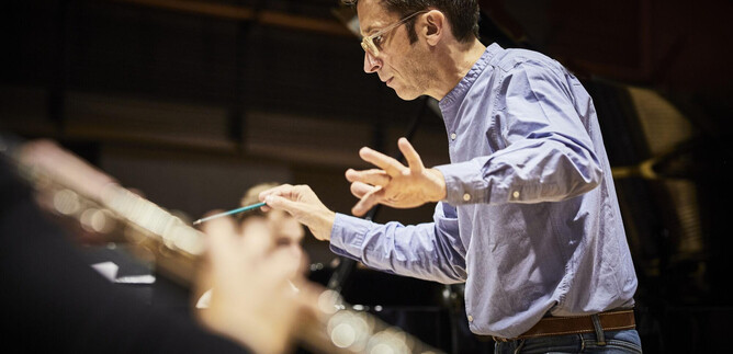 A conductor leads an orchestra with passionate hand gestures. In the foreground, a flute is visible. | © Christophe Raynaud