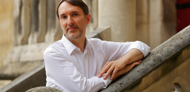 A man in a white shirt is leaning casually against a staircase. In the background, parts of historic buildings can be seen. | © Deyan Parouchev