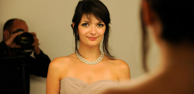 A woman in an elegant dress stands in front of a mirror. She is wearing a necklace and smiling at the camera. | © Luc Jennepin