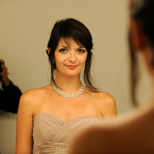 A woman in an elegant dress stands in front of a mirror. She is wearing a necklace and smiling at the camera. | © Luc Jennepin