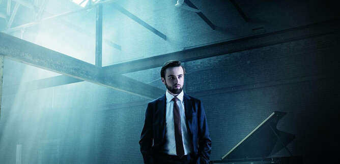 A man in a suit stands in a light-flooded room. In the background, there is a piano. | © Dario Acosta