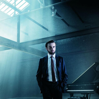 A man in a suit stands in a light-flooded room. In the background, there is a piano. | © Dario Acosta