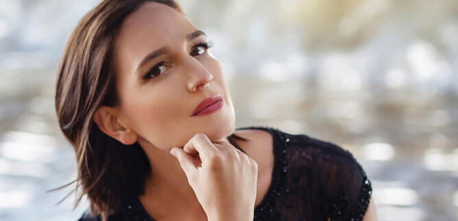 A thoughtful woman with short hair sits in front of a blurred background. She wears a black, sparkly top and has a relaxed expression. | © Yulia Oliver Taylor