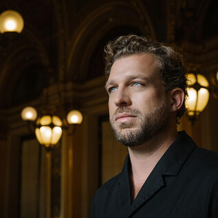 A man with curly hair stands in an elegant room. The background features historical lamps and stylish architecture. | © Chris Gonz