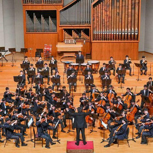 A large orchestra is playing on a stage. The conductor is leading the musicians, who are dressed in elegant suits. | © Konzert Meiji University Nakano
