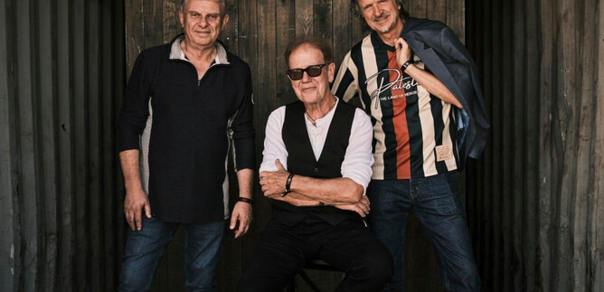 Three men are standing in front of a wooden wall. One is sitting in the middle, while the other two are standing next to him and smiling. | © Weltenklang