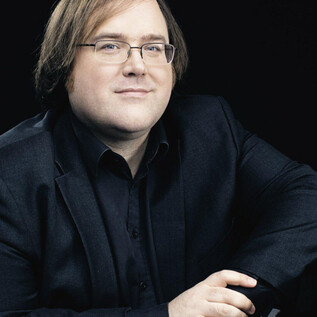 Ein Musiker sitzt entspannt neben einem Klavier. Er trägt einen schwarzen Anzug und schaut freundlich in die Kamera. | © Nikolaj Lund