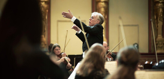 A conductor directs an orchestra in a magnificent hall.  
Musicians from various instrument groups focus on his instructions. | © Meinrad Hofer