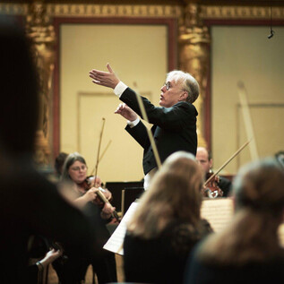 Ein Dirigent leitet ein Orchester in einem prächtigen Saal. Musiker aus verschiedenen Instrumentengruppen konzentrieren sich auf seine Anweisungen. | © Meinrad Hofer