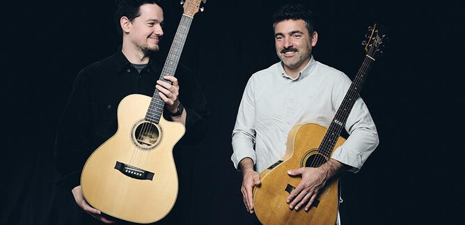 Two men are standing side by side, smiling, holding acoustic guitars. They appear friendly and relaxed against a dark background. | © Elias Maier