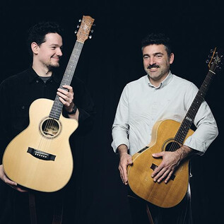 Zwei Männer stehen lächelnd nebeneinander und halten Akustikgitarren. Sie wirken freundlich und entspannt vor einem dunklen Hintergrund. | © Elias Maier
