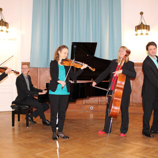 A music group is playing in an elegant room. Four musicians with different instruments are standing together, while a pianist is sitting at the piano. | © Ehrbarsaal