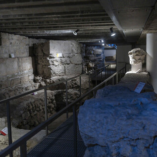 An archaeological excavation site with old stones and walls. There is a metal bridge and illuminated areas for exploration. | © Herbert Rohrer