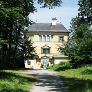 A large, yellow house surrounded by green trees. The path leads directly to the entrance of the building. | © Salzburg Museum