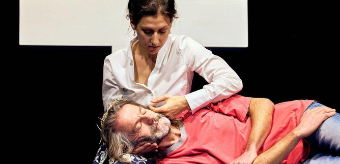 A woman sits next to a man who is lying on a bench. In the background, a large white cross is visible. | © Chris Rogl