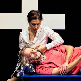 A woman sits next to a man who is lying on a bench. In the background, a large white cross is visible. | © Chris Rogl