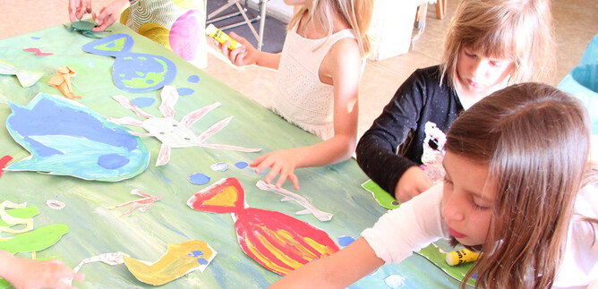 Some children are crafting at a large table with colorful paper cutouts in the shape of sea creatures. They display joy and creativity while working on their art project. | © DomQuartier Salzburg