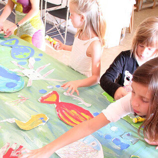 Some children are crafting at a large table with colorful paper cutouts in the shape of sea creatures. They display joy and creativity while working on their art project. | © DomQuartier Salzburg