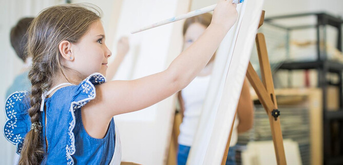 A girl is concentrating on painting on an empty canvas. In the background, other children with easels can be seen. | © DomQuartier Salzburg
