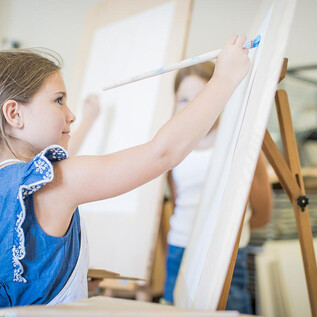 A girl is concentrating on painting on an empty canvas. In the background, other children with easels can be seen. | © DomQuartier Salzburg