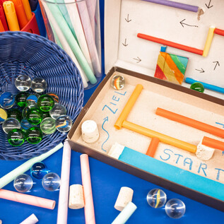 Colorful crafting materials are spread out on a wooden table. There are pieces of paper, scissors, and craft sticks in various colors visible. | © Salzburg Museum/Maurice Rigaud