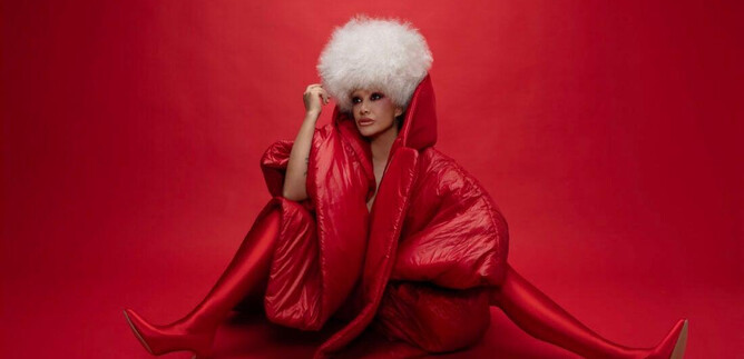 A woman sits against a red background, dressed in a striking red coat and high red shoes. She wears a large white fur hat and poses elegantly. | © Studio1538