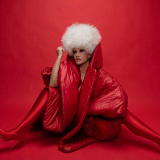 Eine Frau sitzt vor einem roten Hintergrund, gekleidet in einen auffälligen roten Mantel und hohe rote Schuhe. Sie trägt eine große, weiße Fellmütze und posiert elegant. | © Studio1538