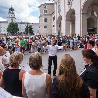 A large crowd gathers in a square to listen to a choir performance. In the foreground, a conductor leads the singers. | © Chorverband Salzburg/Moser