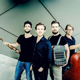 A four-member music group stands in front of a modern, geometric wall. Each musician holds an instrument, including a violin, cello, and another more specialized music machine. | © Andrej Grilc