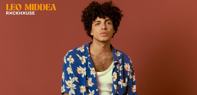 A young man with curly hair is wearing a colorful, floral shirt. The background is in a warm color tone. | © Elisa Maciel