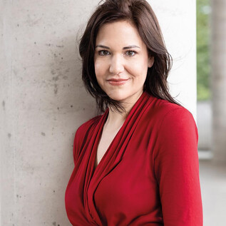 A woman in a black suit with curly hair stands smiling in front of an orange wall. She has her arms crossed and looks friendly. | © Alexandra Grill