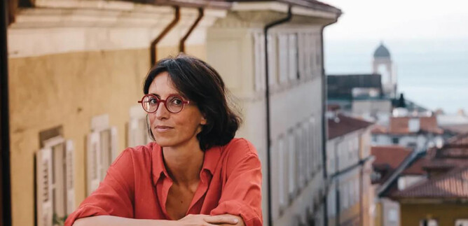 A woman in a black suit with curly hair stands smiling in front of an orange wall. She has her arms crossed and looks friendly. | © Adolfo Frediani