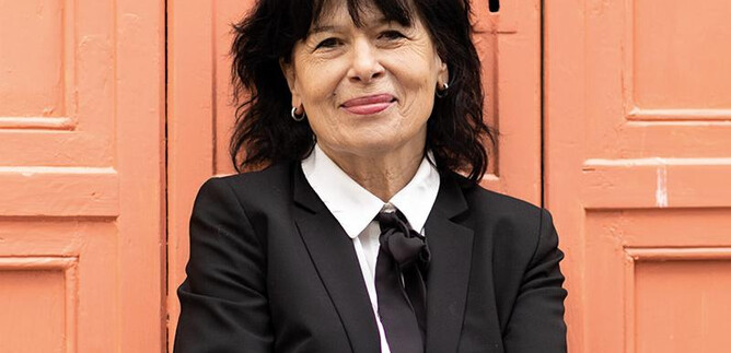 A woman in a black suit with curly hair stands smiling in front of an orange wall. She has her arms crossed and looks friendly. | © Minitta Kandlbauer