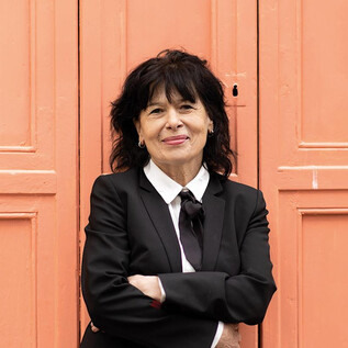 A woman in a black suit with curly hair stands smiling in front of an orange wall. She has her arms crossed and looks friendly. | © Minitta Kandlbauer