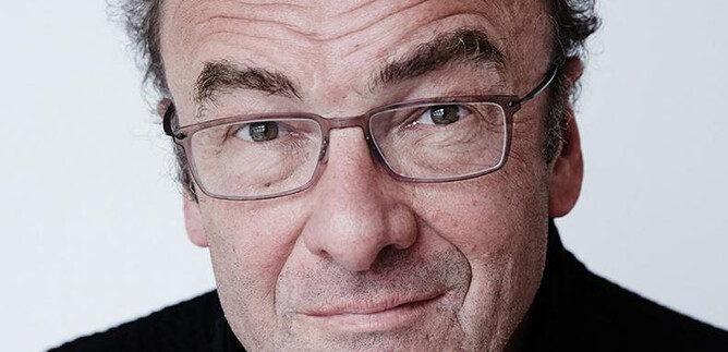 A woman in a black suit with curly hair stands smiling in front of an orange wall. She has her arms crossed and looks friendly. | © Rafaela Proell