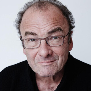 A woman in a black suit with curly hair stands smiling in front of an orange wall. She has her arms crossed and looks friendly. | © Rafaela Proell
