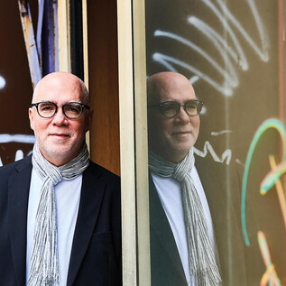 A woman in a black suit with curly hair stands smiling in front of an orange wall. She has her arms crossed and looks friendly. | © Aleksandra Pawloff