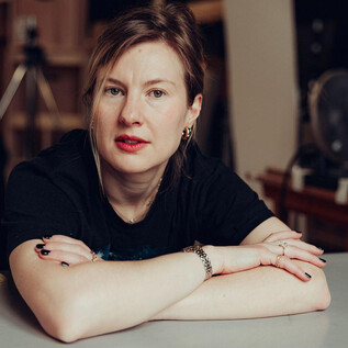 Žena sedí u stolu a hledí přímo do kamery. Má na sobě černé tričko a má zamyšlený výraz. | © Joel Heyd