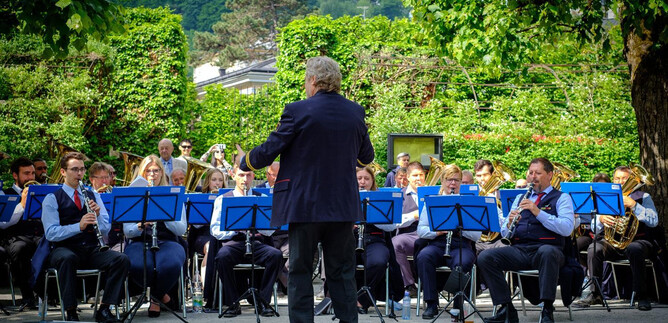 Ein bläserisches Ensemble spielt im Freien. Der Dirigent steht vor der Gruppe, umgeben von grünen Bäumen. | © knaro.at
