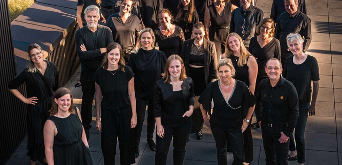 Skupina lidí stojí v moderním, městském prostředí. Všichni nosí černé oblečení a usmívají se do kamery. | © Vokalensemble QuasiSolo e.V.