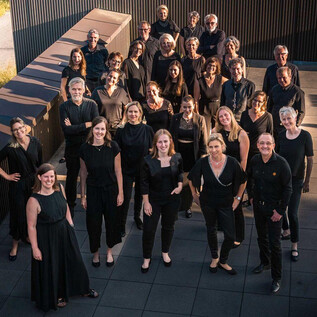 A group of people stands in a modern, urban environment. They are all wearing black clothing and smiling at the camera. | © Vokalensemble QuasiSolo e.V.